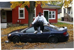 Darryll Stinson sitting on a damaged car in a residential neighborhood, representing resilience after adversity