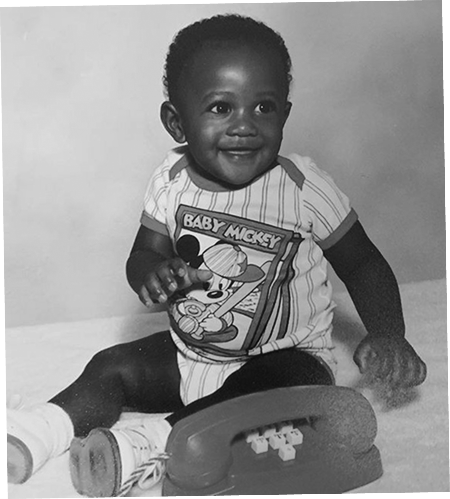 Baby photo of Darryll Stinson sitting on the floor, smiling and wearing a Mickey Mouse outfit
